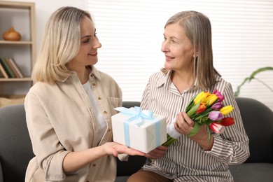 Photo of Smiling daughter congratulating her mom with bouquet of tulips and gift on sofa at home. Happy Mother's Day