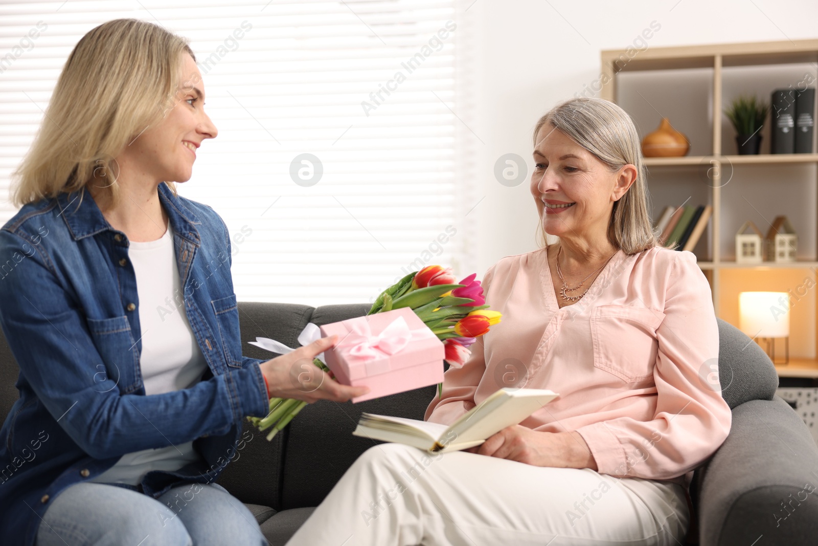 Photo of Smiling daughter congratulating her mom with bouquet of tulips and gift on sofa at home. Happy Mother's Day