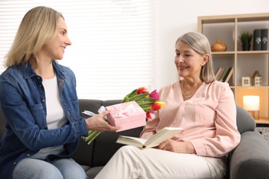 Photo of Smiling daughter congratulating her mom with bouquet of tulips and gift on sofa at home. Happy Mother's Day