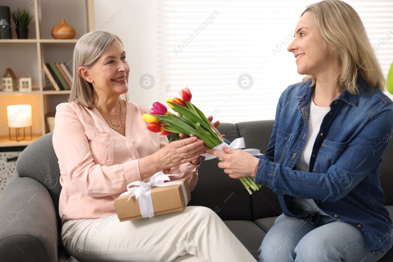 Photo of Smiling daughter congratulating her mom with bouquet of tulips and gift on sofa at home. Happy Mother's Day