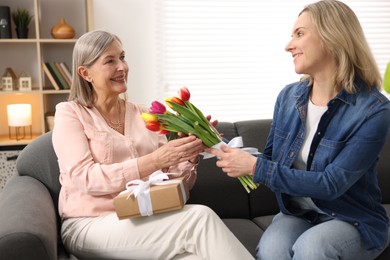 Photo of Smiling daughter congratulating her mom with bouquet of tulips and gift on sofa at home. Happy Mother's Day