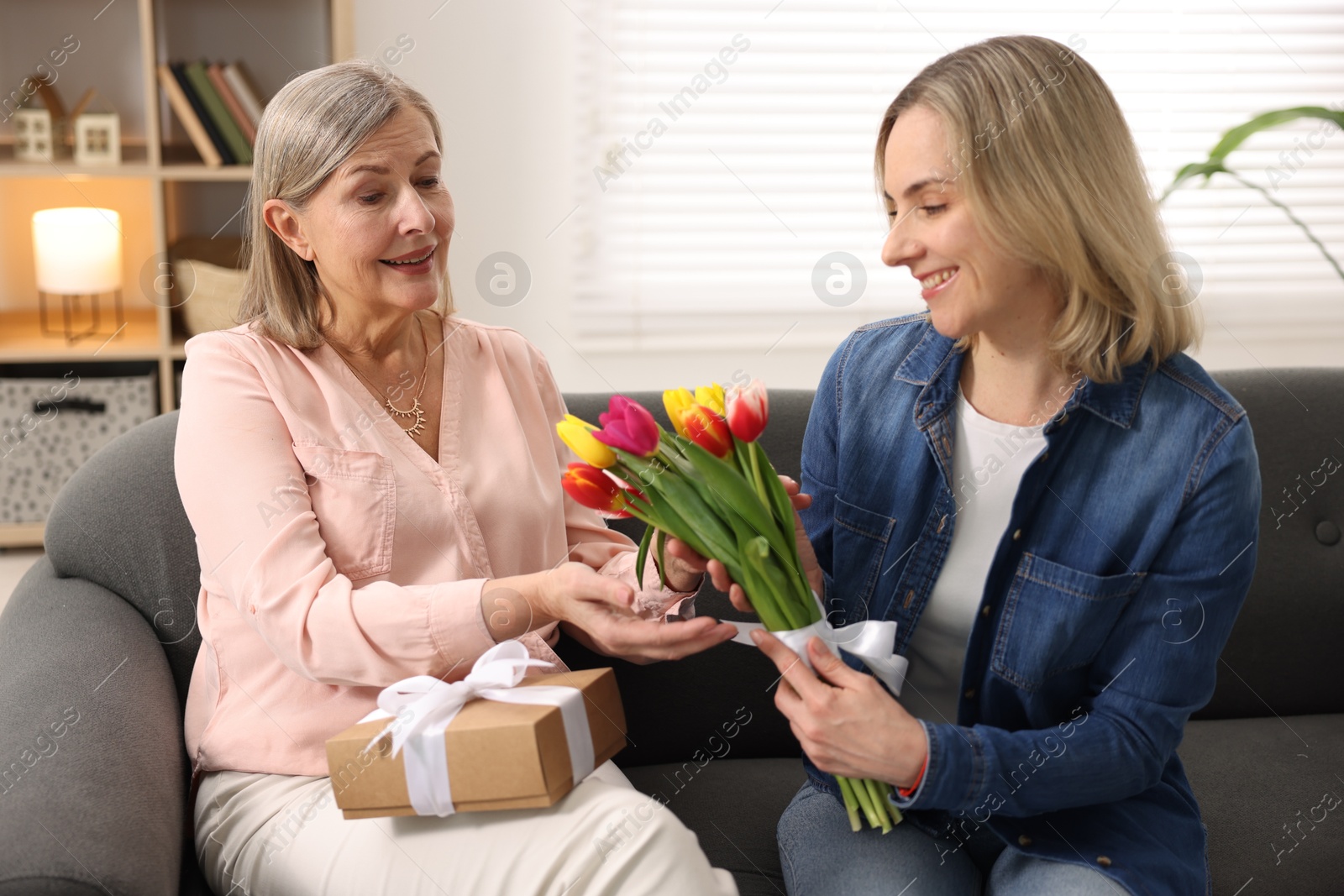 Photo of Smiling daughter congratulating her mom with bouquet of tulips and gift on sofa at home. Happy Mother's Day