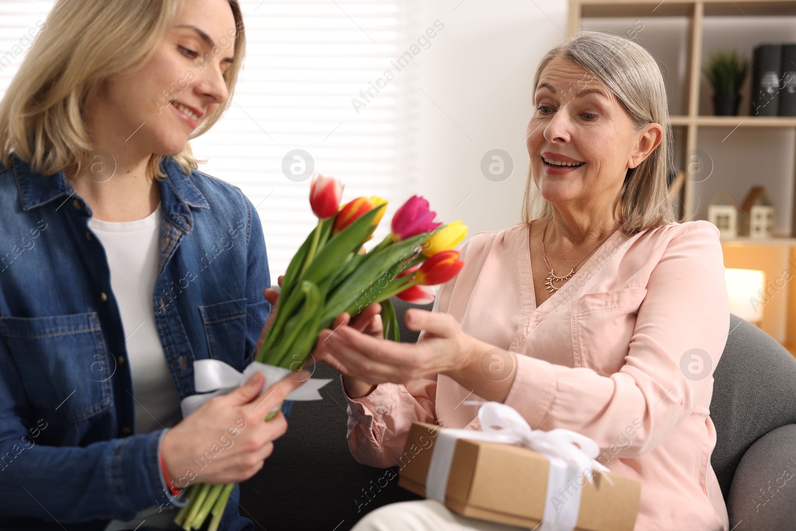 Photo of Smiling daughter congratulating her mom with bouquet of tulips and gift on sofa at home. Happy Mother's Day