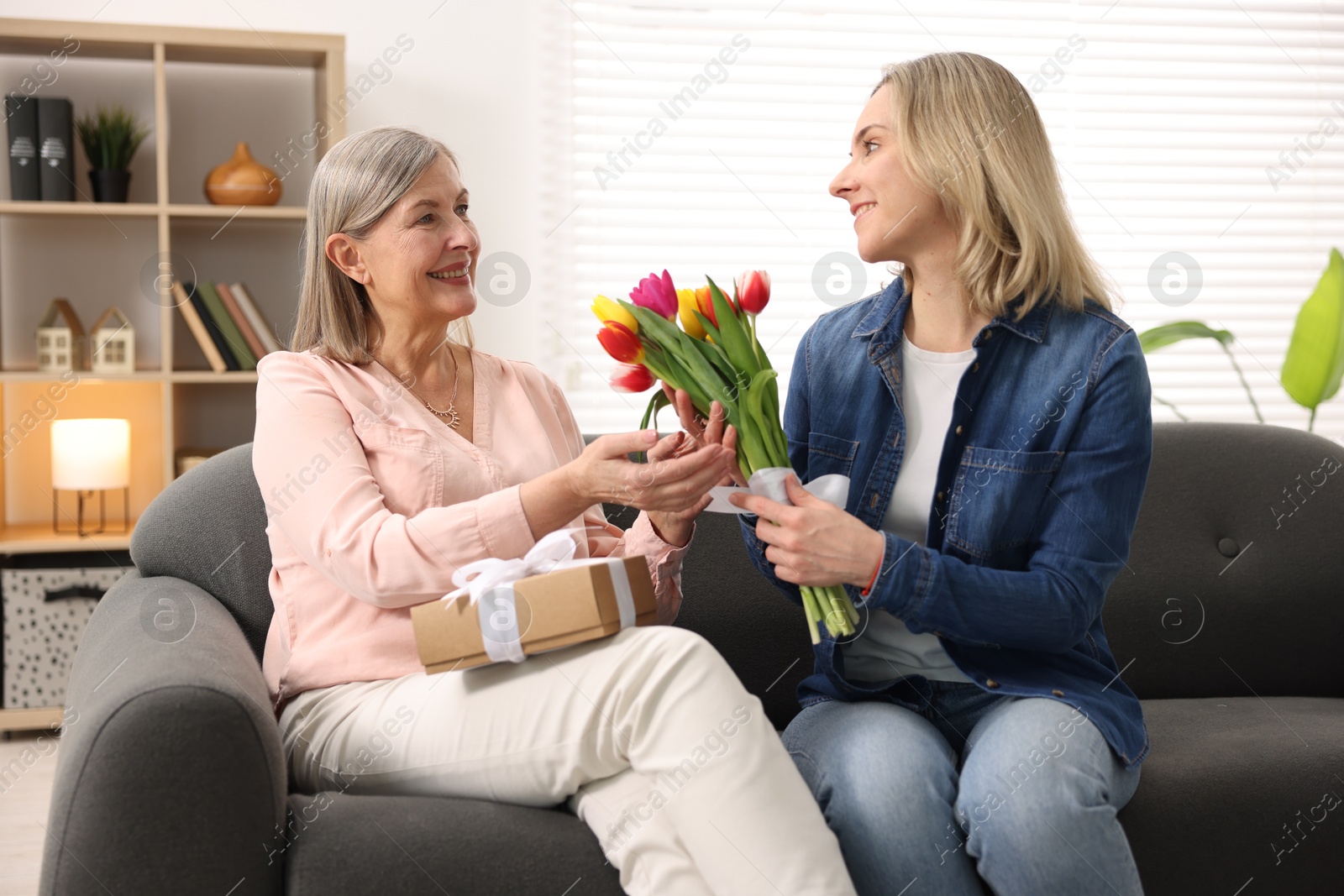 Photo of Smiling daughter congratulating her mom with bouquet of tulips and gift on sofa at home. Happy Mother's Day