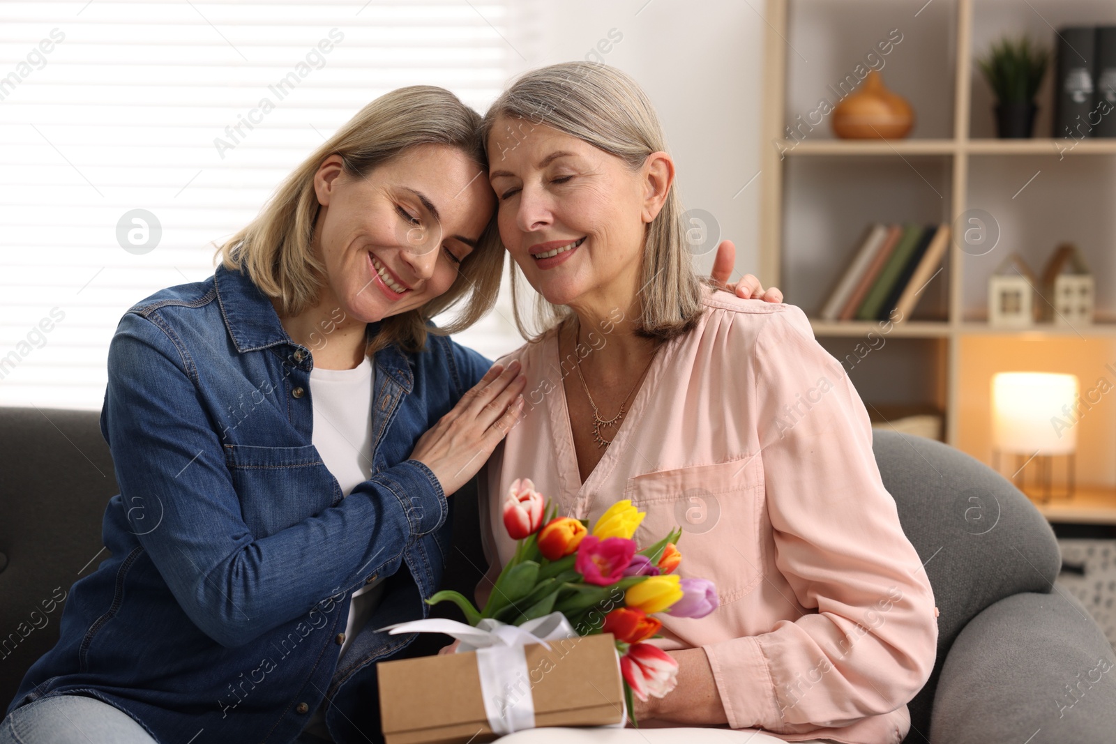 Photo of Smiling woman with bouquet of tulips, gift and her daughter on sofa at home. Happy Mother's Day