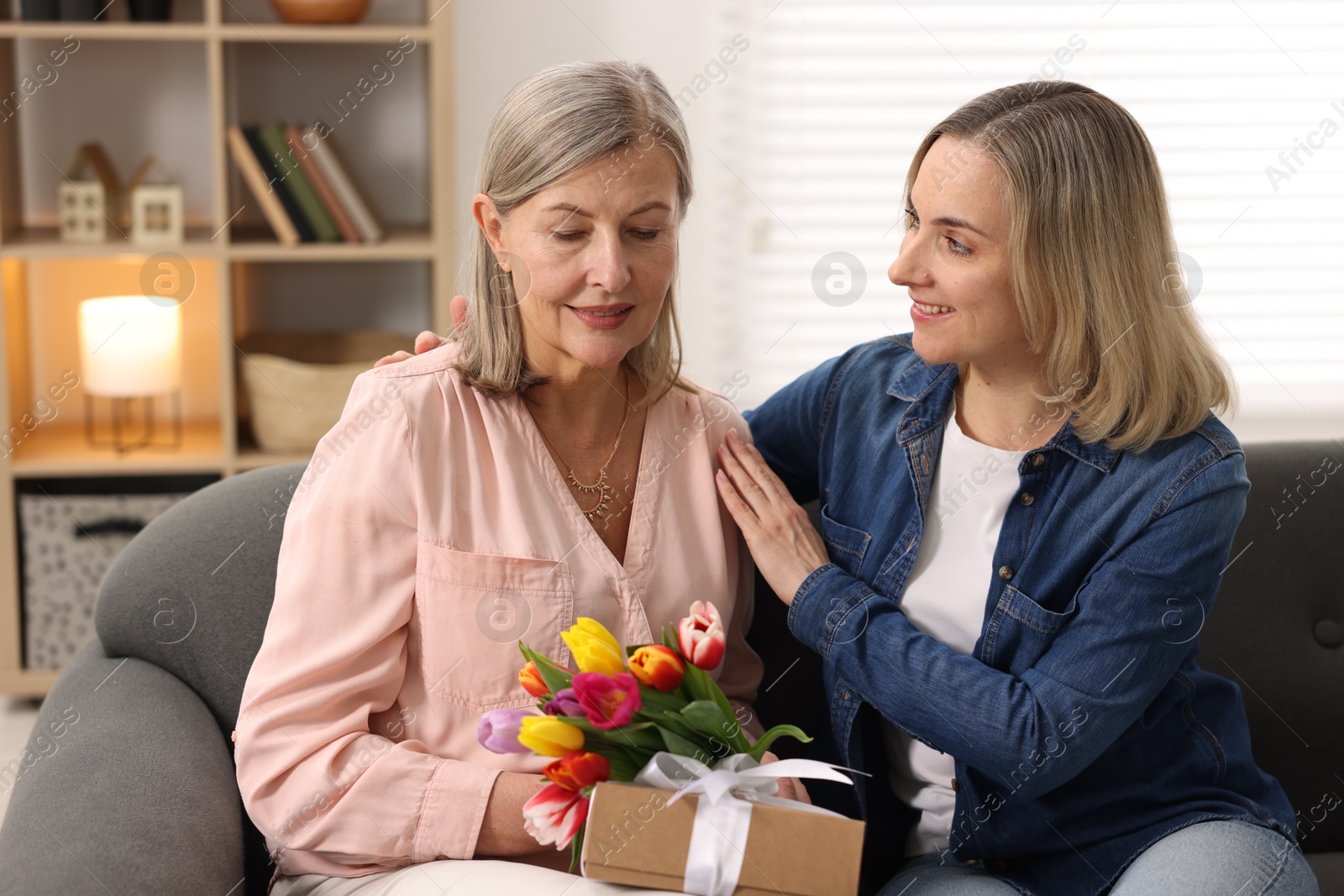 Photo of Smiling woman with bouquet of tulips, gift and her daughter on sofa at home. Happy Mother's Day