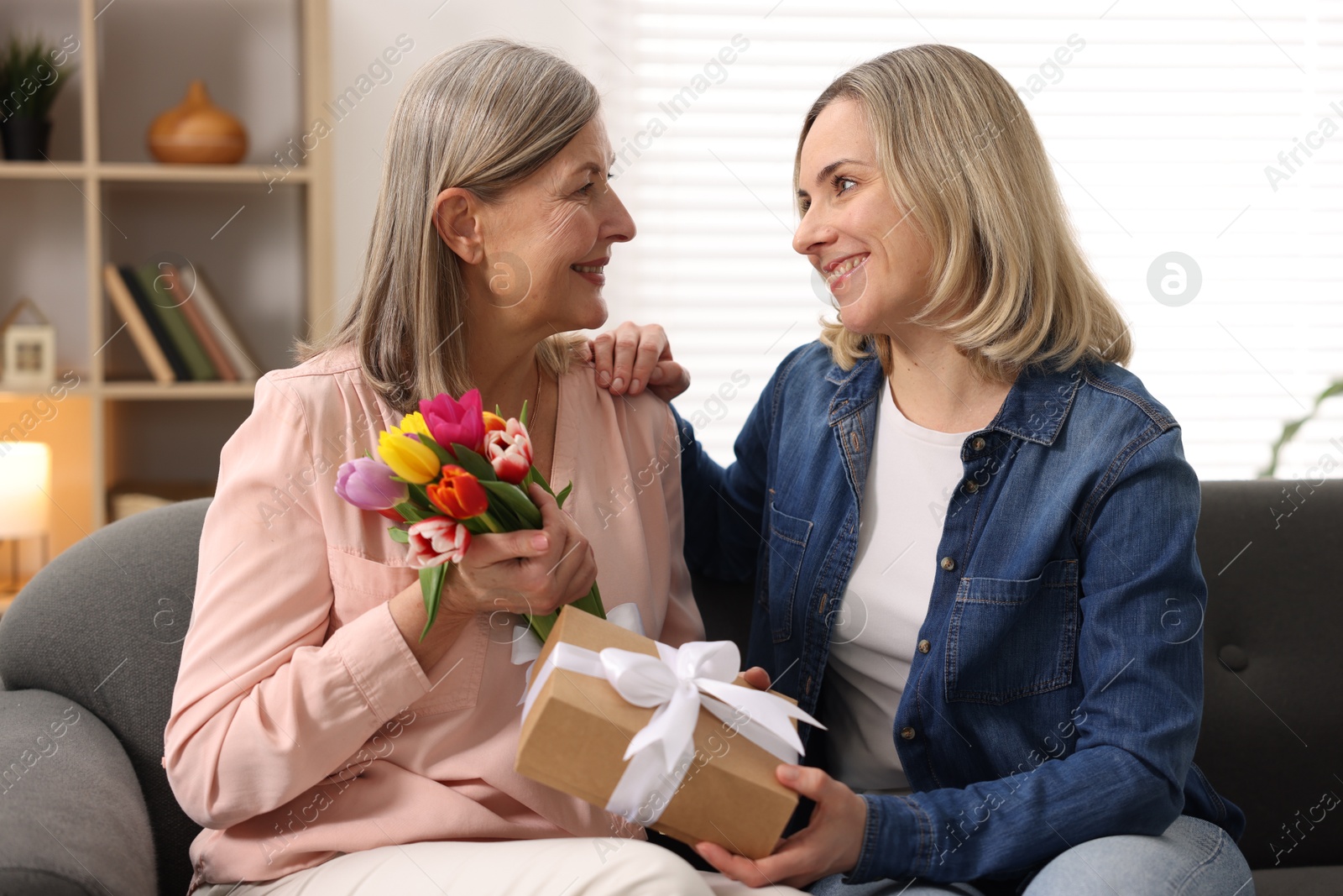 Photo of Smiling daughter congratulating her mom with bouquet of tulips and gift on sofa at home. Happy Mother's Day