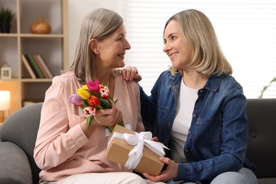 Photo of Smiling daughter congratulating her mom with bouquet of tulips and gift on sofa at home. Happy Mother's Day