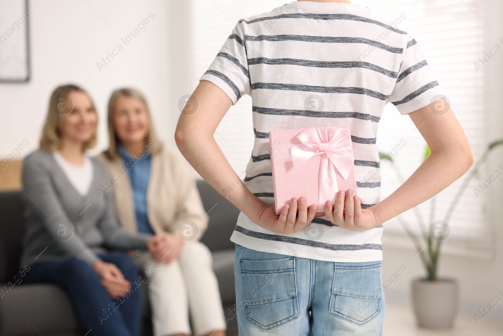 Photo of Boy hiding gift box behind back for his parents at home, selective focus. Happy Mother's Day