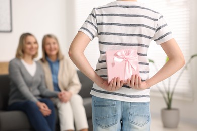 Boy hiding gift box behind back for his parents at home, selective focus. Happy Mother's Day