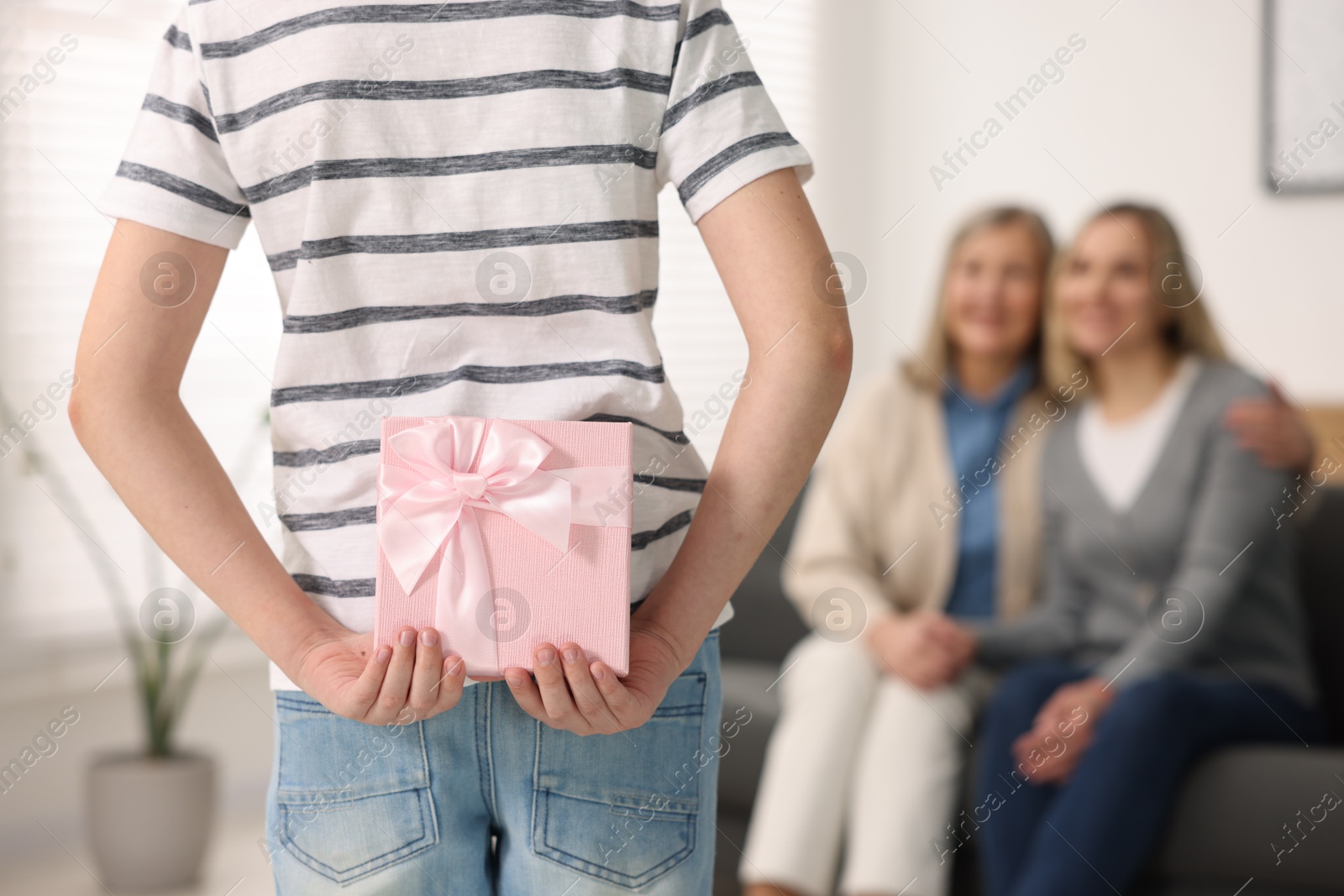 Photo of Boy hiding gift box behind back for his parents at home, selective focus. Happy Mother's Day