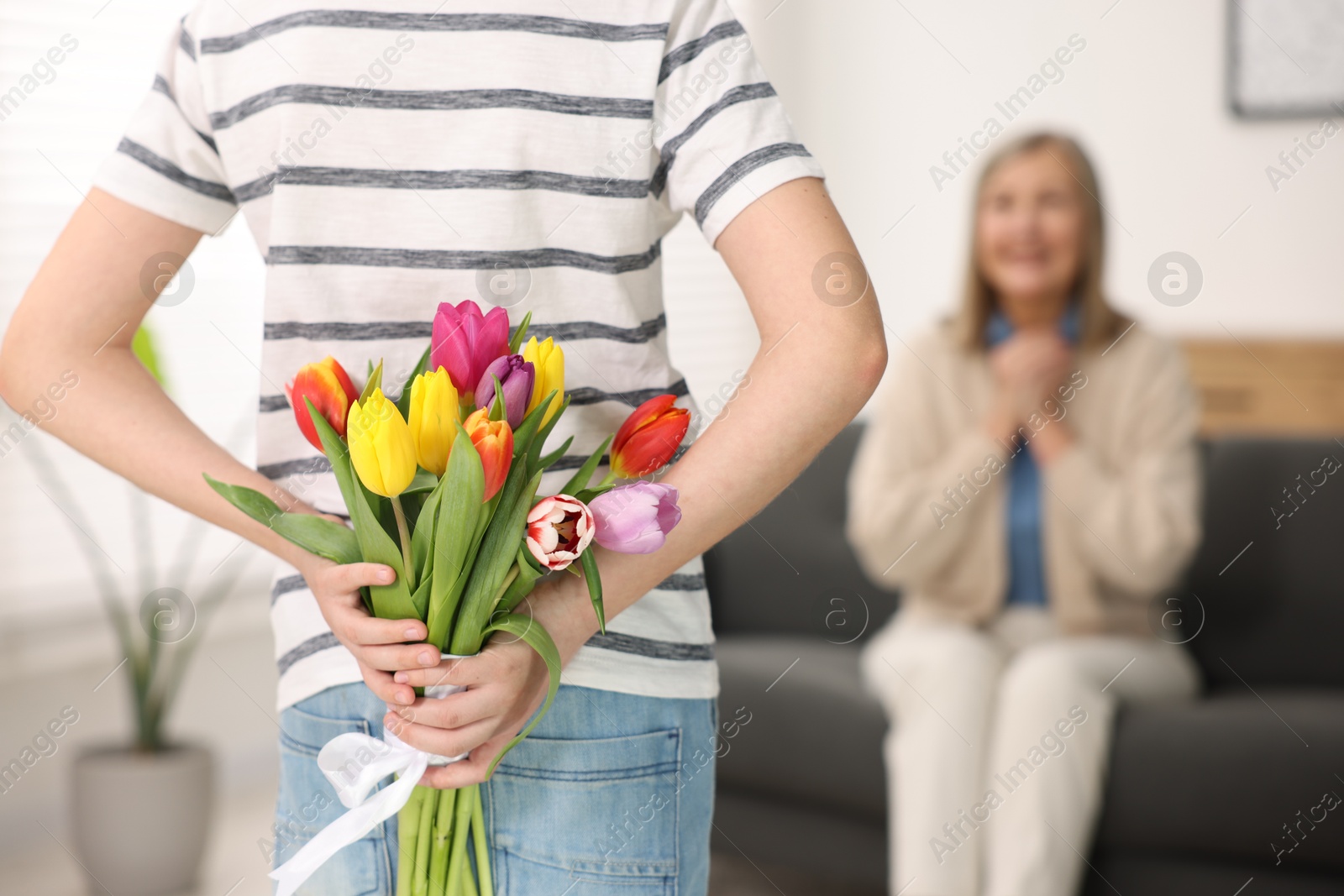 Photo of Boy hiding bouquet of tulips behind back for his mom at home, selective focus. Happy Mother's Day