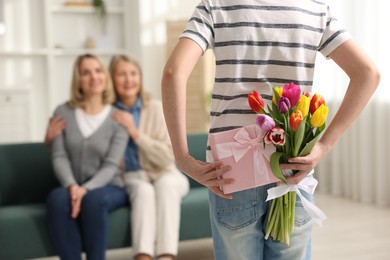 Photo of Boy hiding bouquet of tulips and gift box behind back for his parents at home, selective focus. Happy Mother's Day