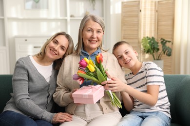 Photo of Smiling woman with bouquet of tulips, gift and her relatives on sofa at home. Happy Mother's Day