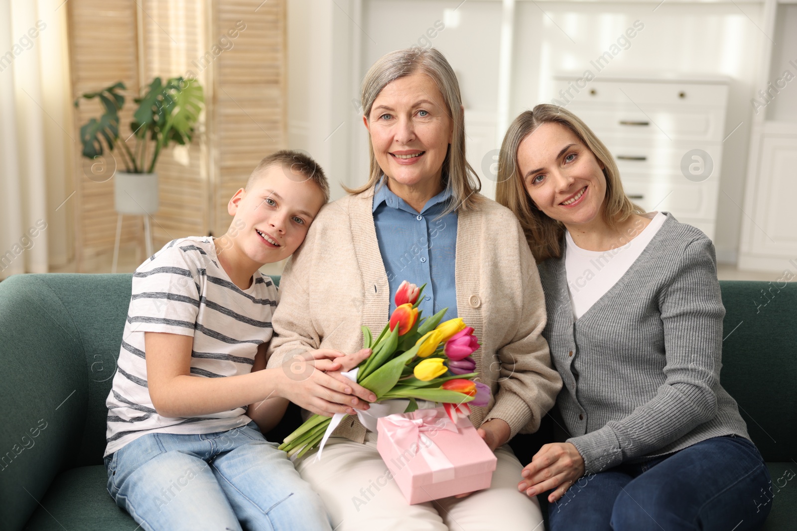 Photo of Smiling woman with bouquet of tulips, gift and her relatives on sofa at home. Happy Mother's Day