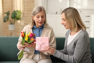 Smiling daughter congratulating her mom with bouquet of tulips and gift on sofa at home. Happy Mother's Day