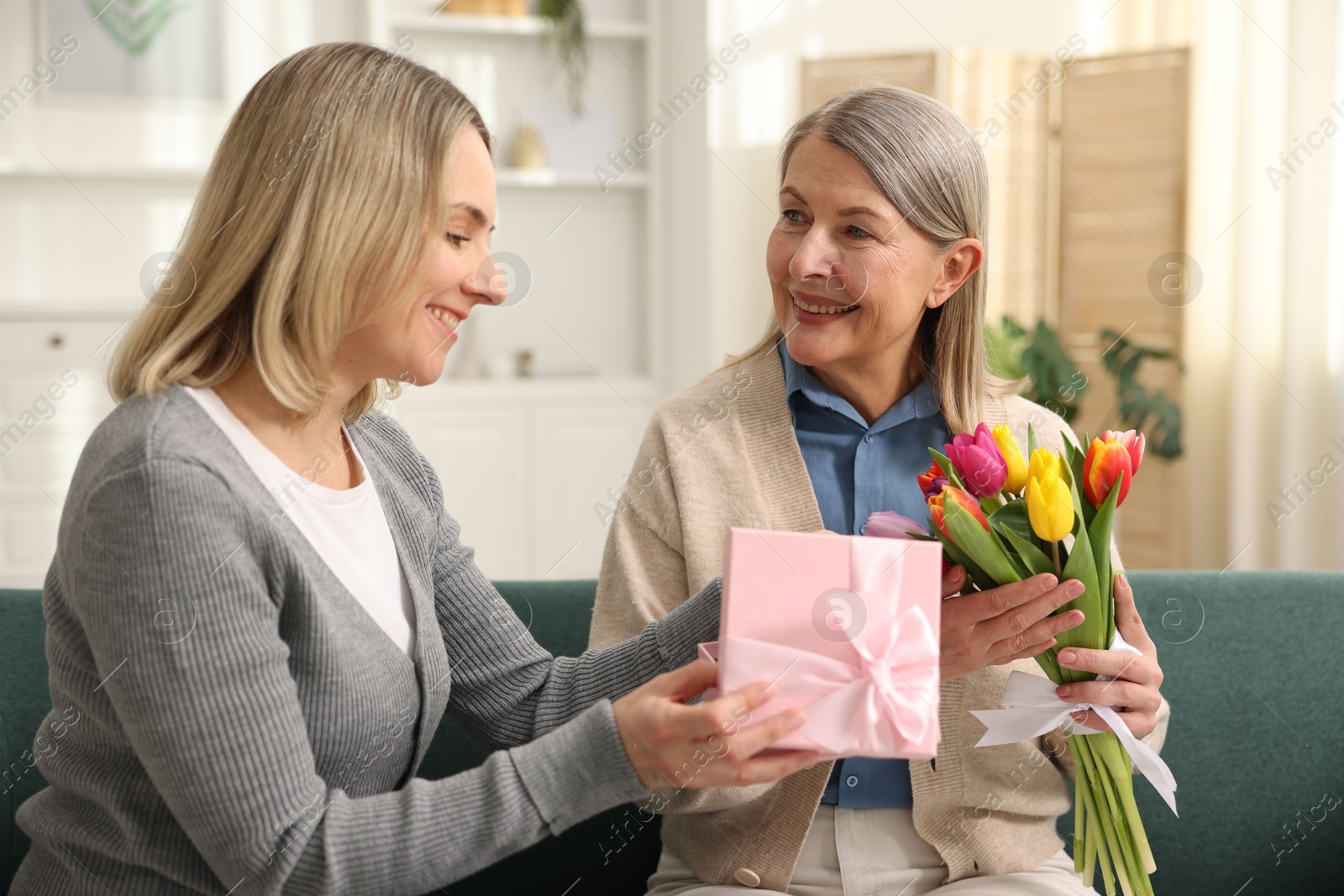 Photo of Smiling daughter congratulating her mom with bouquet of tulips and gift on sofa at home. Happy Mother's Day