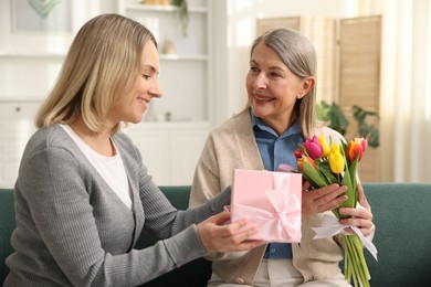 Photo of Smiling daughter congratulating her mom with bouquet of tulips and gift on sofa at home. Happy Mother's Day