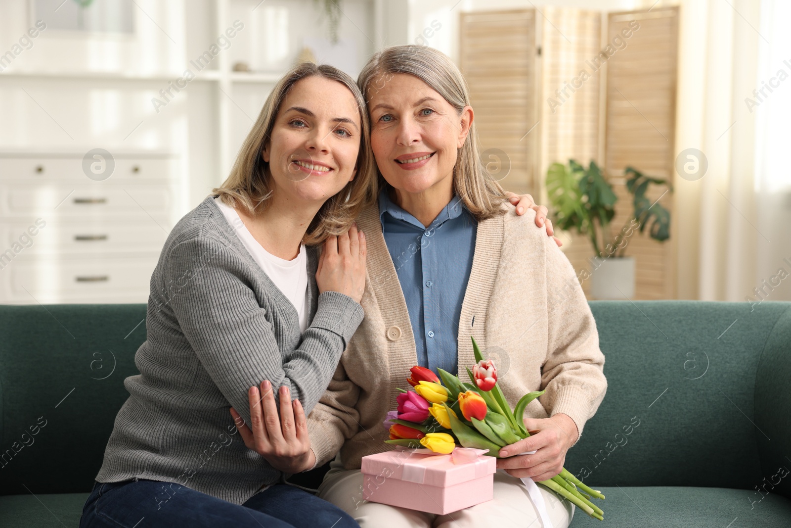 Photo of Smiling woman with bouquet of tulips, gift and her daughter on sofa at home. Happy Mother's Day