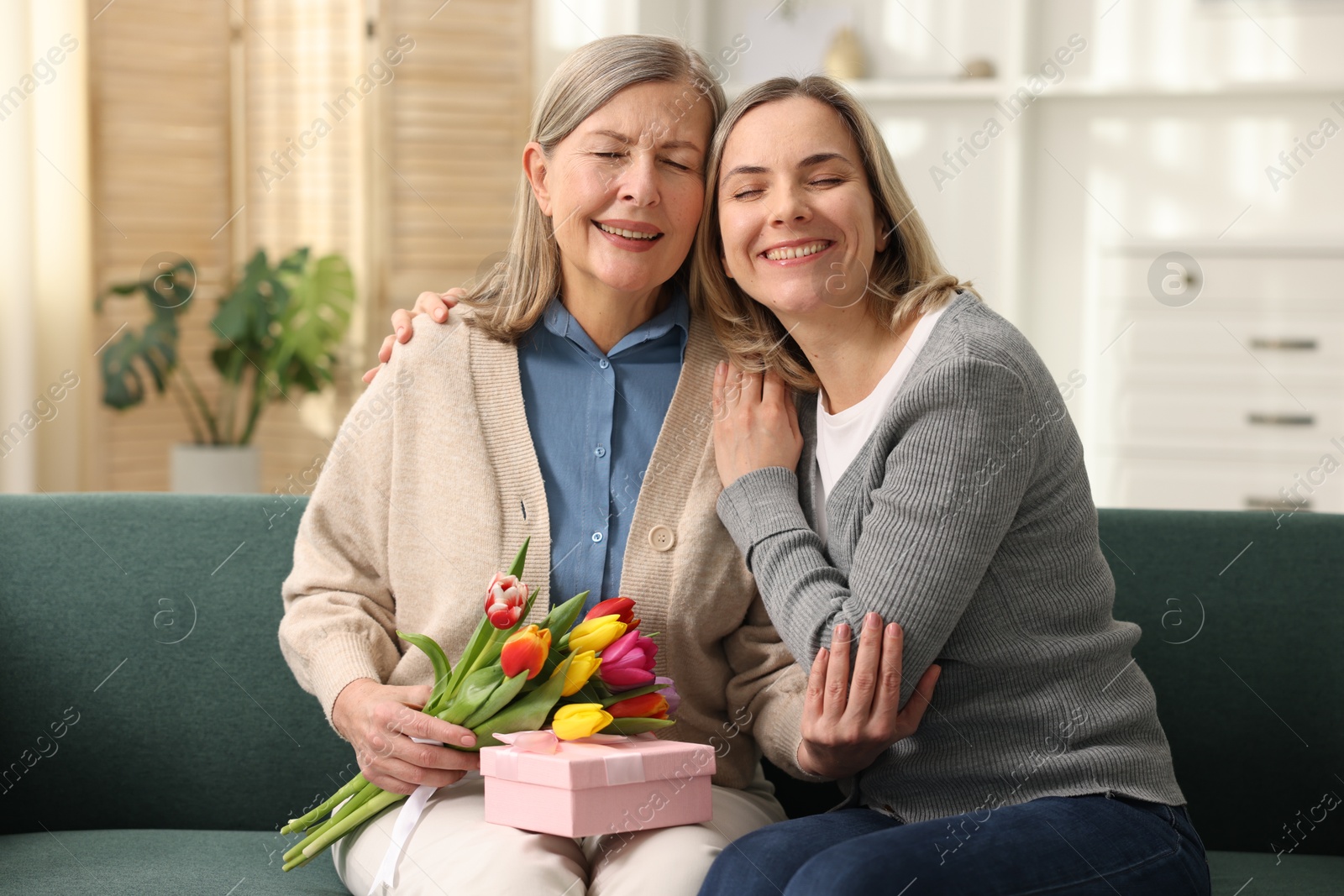 Photo of Smiling daughter congratulating her mom with bouquet of tulips and gift on sofa at home. Happy Mother's Day