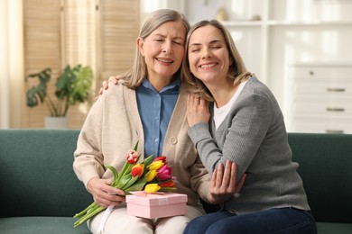 Smiling daughter congratulating her mom with bouquet of tulips and gift on sofa at home. Happy Mother's Day