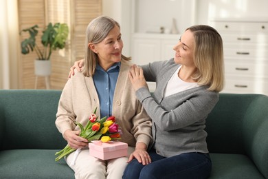 Photo of Smiling daughter congratulating her mom with bouquet of tulips and gift on sofa at home. Happy Mother's Day