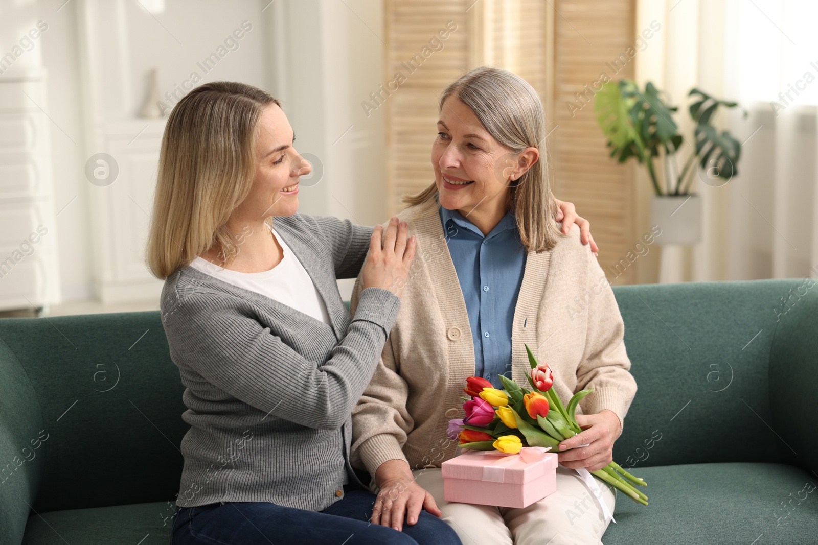 Photo of Smiling woman with bouquet of tulips, gift and her daughter on sofa at home. Happy Mother's Day