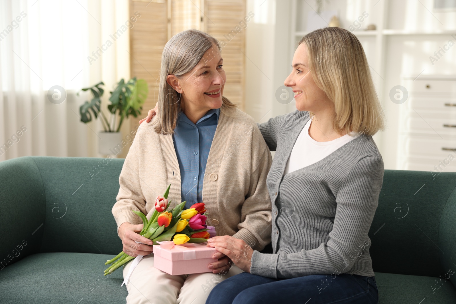 Photo of Smiling woman with bouquet of tulips, gift and her daughter on sofa at home. Happy Mother's Day