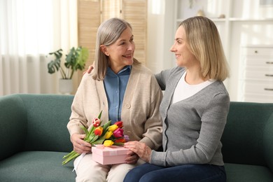 Smiling woman with bouquet of tulips, gift and her daughter on sofa at home. Happy Mother's Day