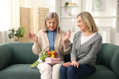 Photo of Smiling daughter congratulating her mom with bouquet of tulips and gift on sofa at home. Happy Mother's Day