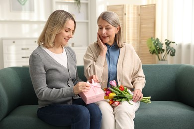 Photo of Smiling daughter congratulating her mom with bouquet of tulips and gift on sofa at home. Happy Mother's Day