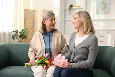 Photo of Smiling daughter congratulating her mom with bouquet of tulips and gift on sofa at home. Happy Mother's Day