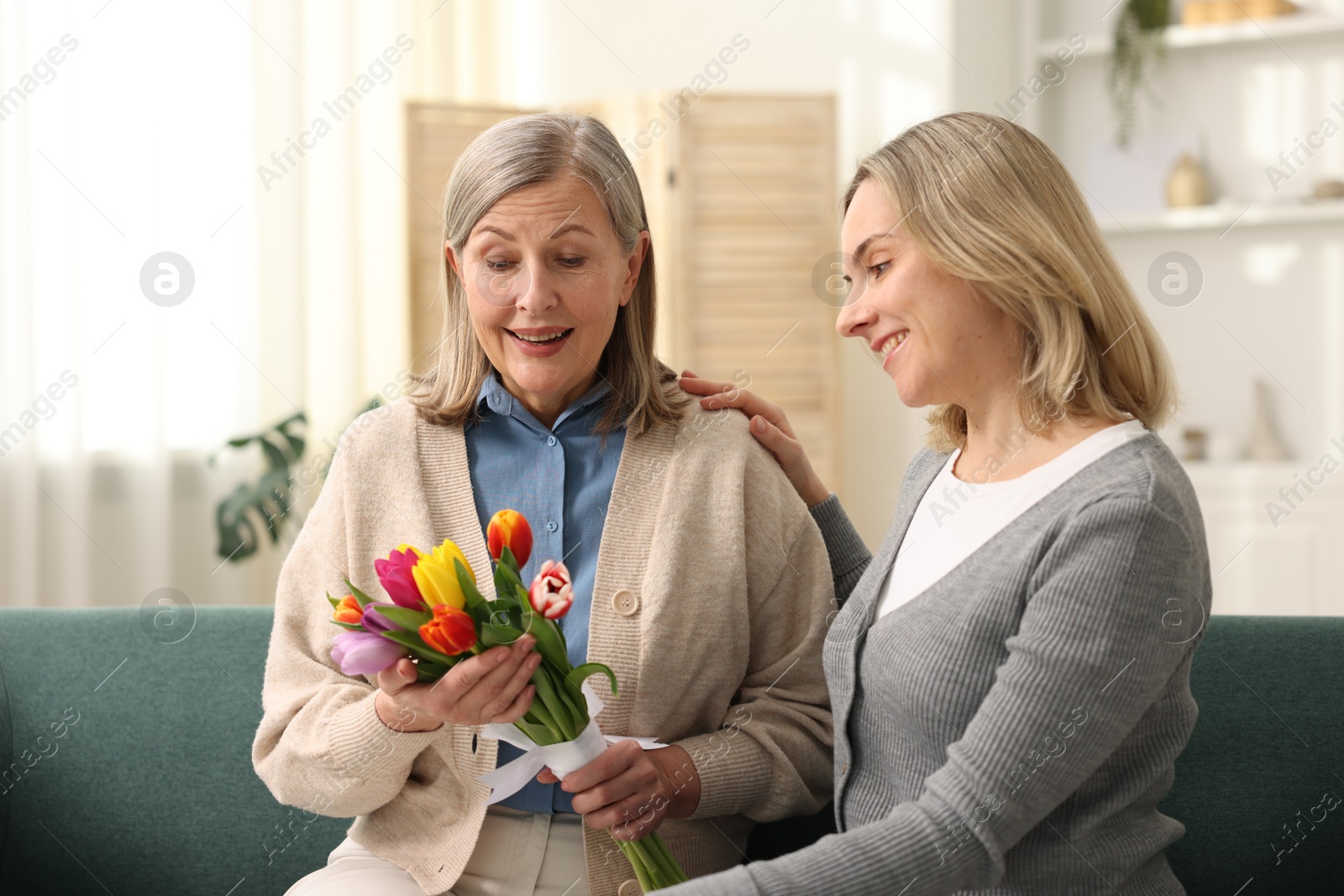 Photo of Smiling woman with bouquet of tulips and her daughter on sofa at home. Happy Mother's Day