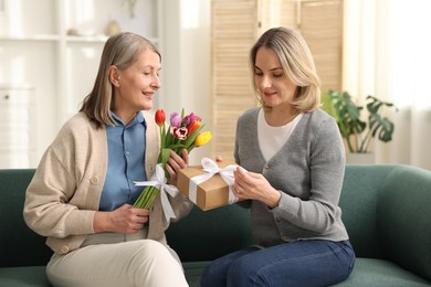 Photo of Smiling daughter congratulating her mom with bouquet of tulips and gift on sofa at home. Happy Mother's Day