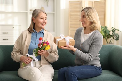 Photo of Smiling daughter congratulating her mom with bouquet of tulips and gift on sofa at home. Happy Mother's Day