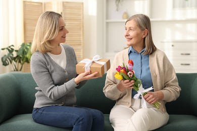 Photo of Smiling daughter congratulating her mom with bouquet of tulips and gift on sofa at home. Happy Mother's Day