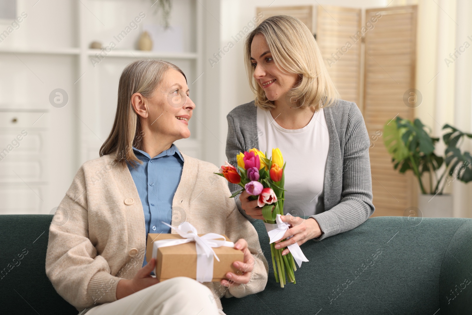 Photo of Smiling daughter congratulating her mom with bouquet of tulips and gift at home. Happy Mother's Day