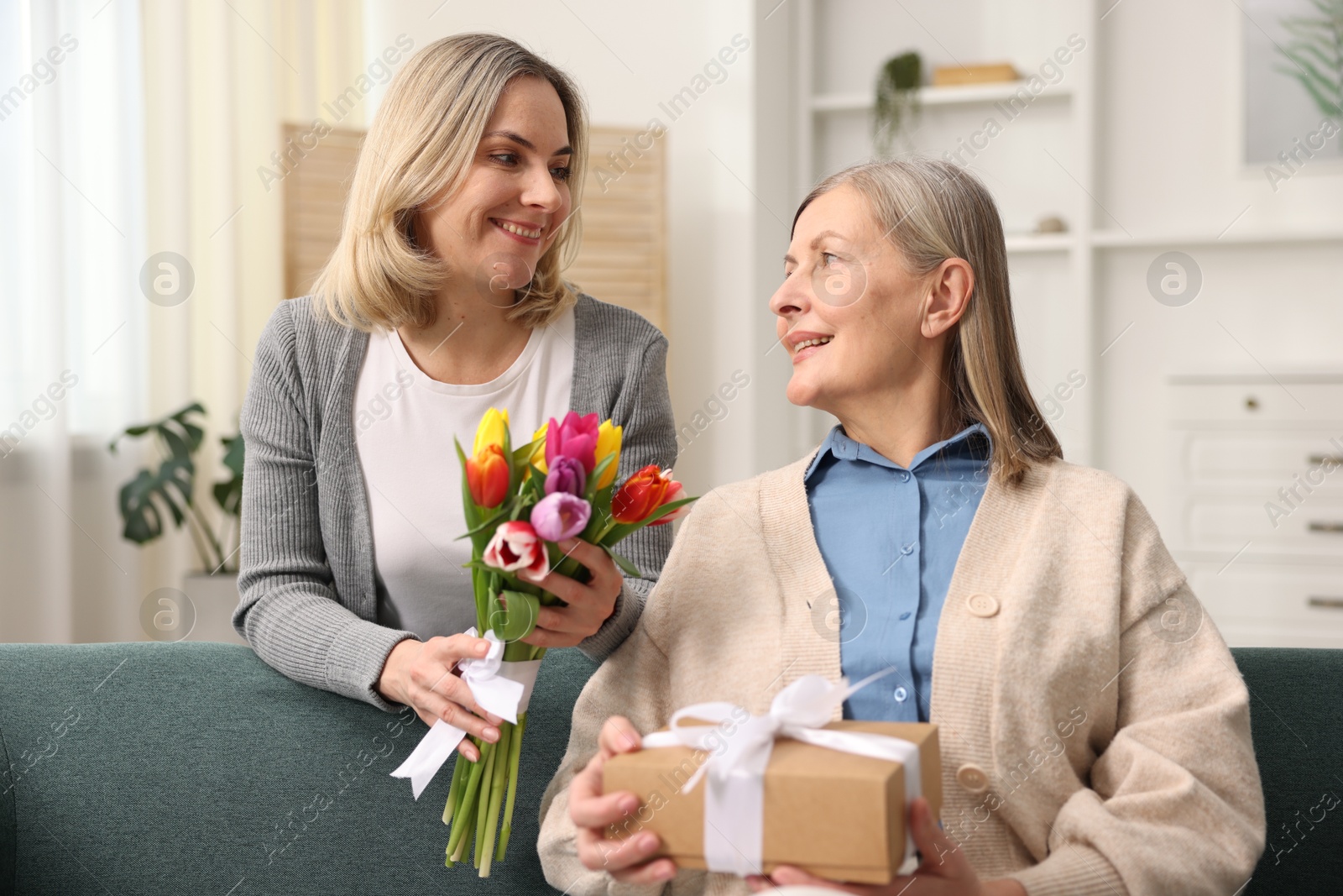 Photo of Smiling daughter congratulating her mom with bouquet of tulips and gift at home. Happy Mother's Day