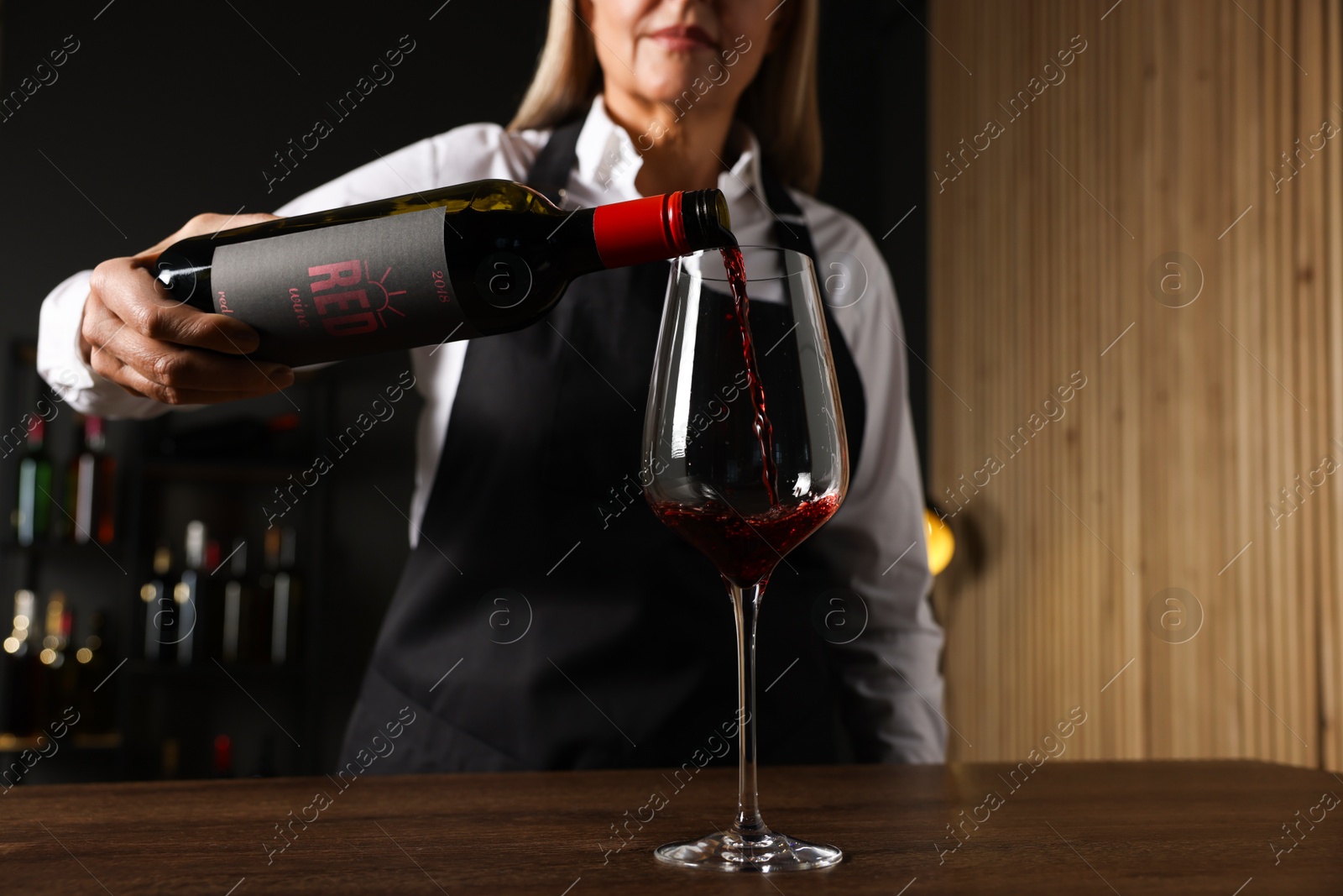 Photo of Professional sommelier pouring red wine into glass at wooden table indoors, closeup