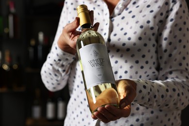 Photo of Professional sommelier holding bottle of white wine indoors, closeup