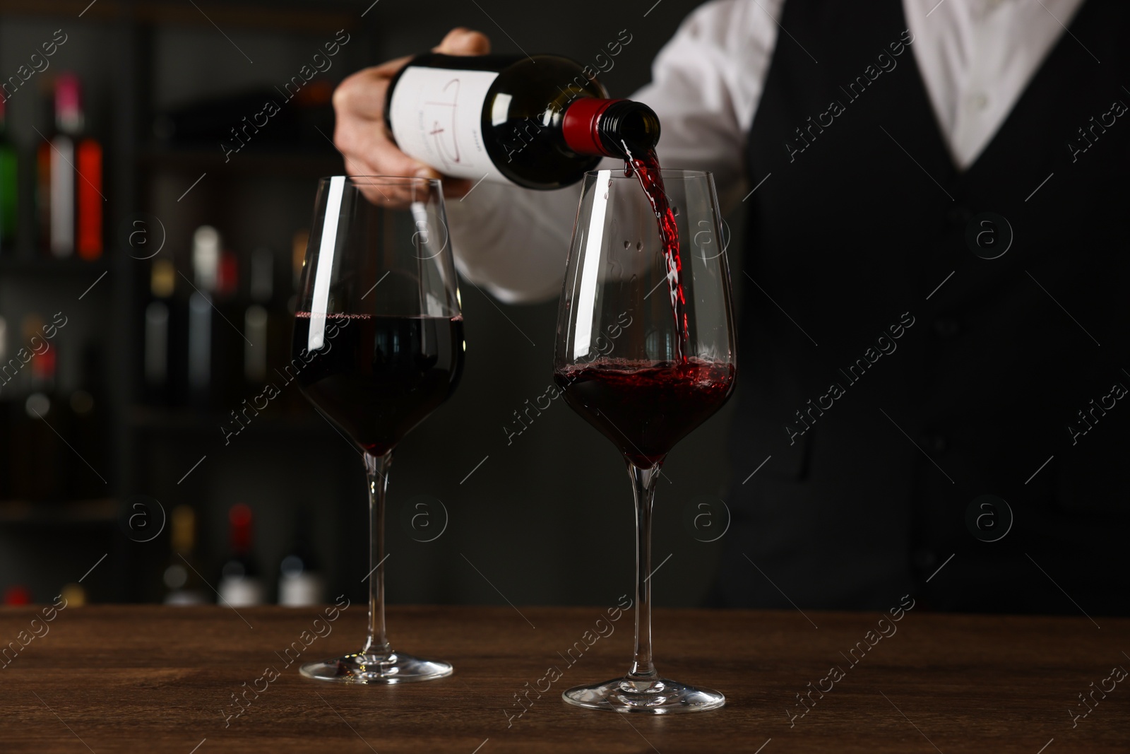 Photo of Professional sommelier pouring red wine into glasses at wooden table indoors, closeup