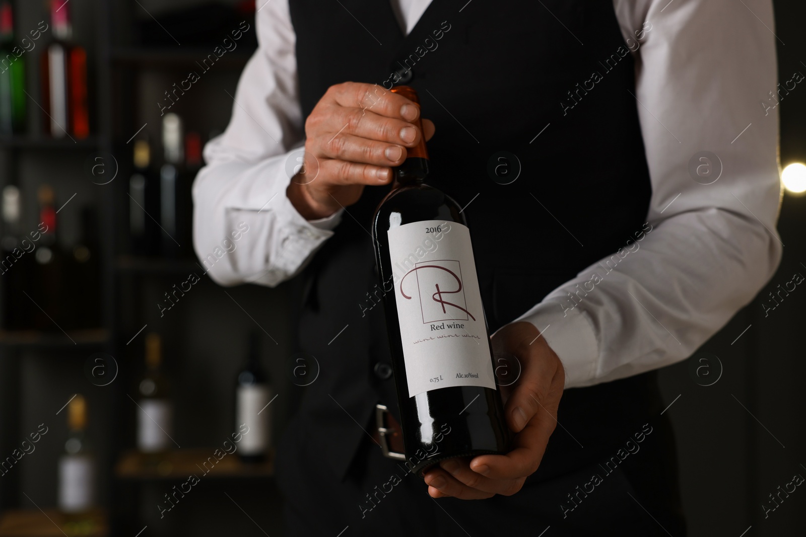 Photo of Professional sommelier holding bottle of red wine indoors, closeup