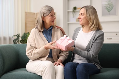 Photo of Smiling daughter congratulating her mom with gift on sofa at home. Happy Mother's Day