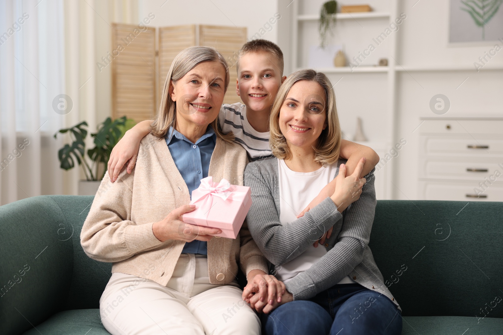 Photo of Family portrait of smiling woman with gift box and her relatives on sofa at home. Happy Mother's Day