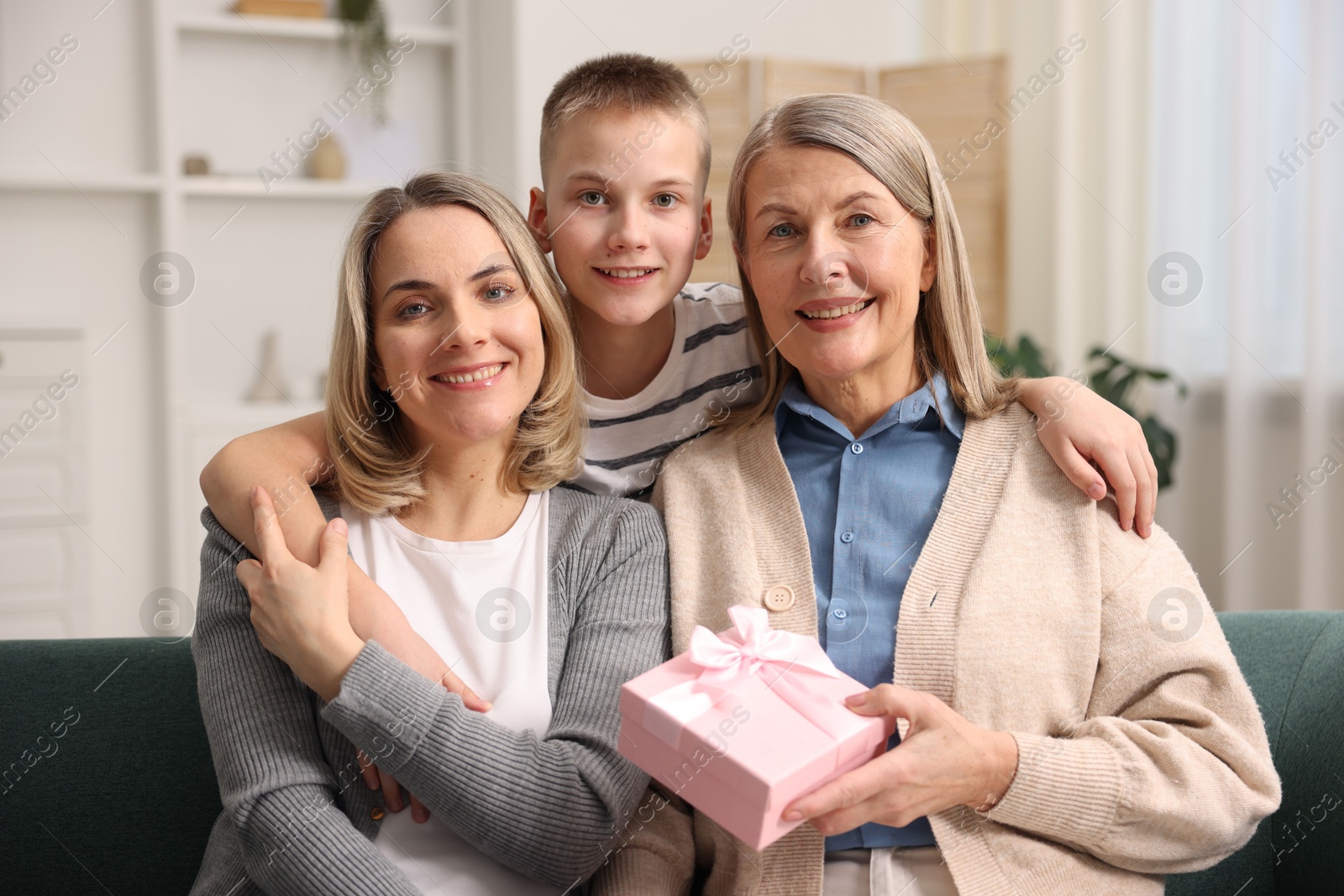 Photo of Family portrait of smiling woman with gift box and her relatives on sofa at home. Happy Mother's Day