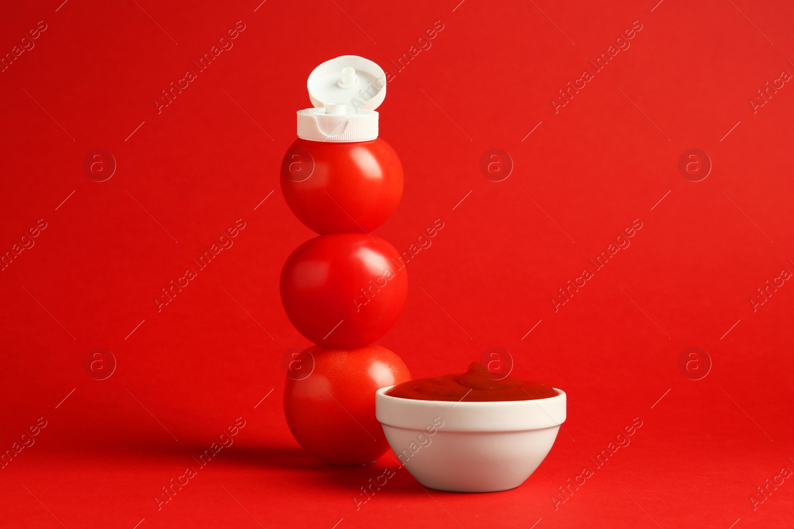 Photo of Stack of fresh tomatoes with plastic cap as bottle and ketchup in bowl on red background