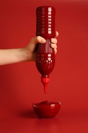 Photo of Woman squeezing ketchup from bottle into bowl on red background, closeup