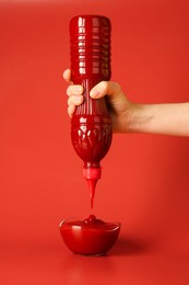 Photo of Woman squeezing ketchup from bottle into bowl on red background, closeup