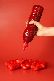 Photo of Woman squeezing ketchup from bottle over tomatoes on red background, closeup