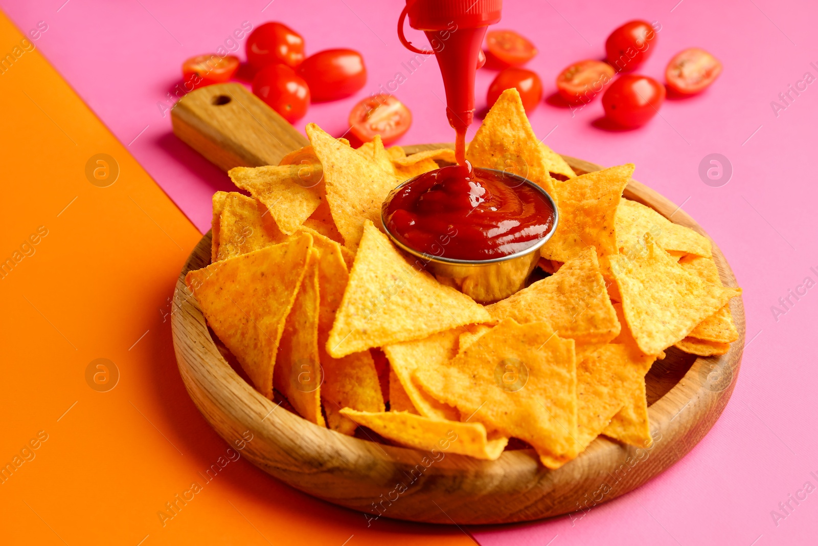 Photo of Pouring tasty ketchup in bowl with nachos on pink background, closeup